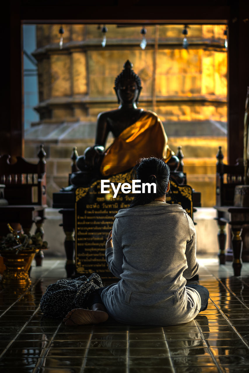 Rear view of woman sitting on floor against statue in temple