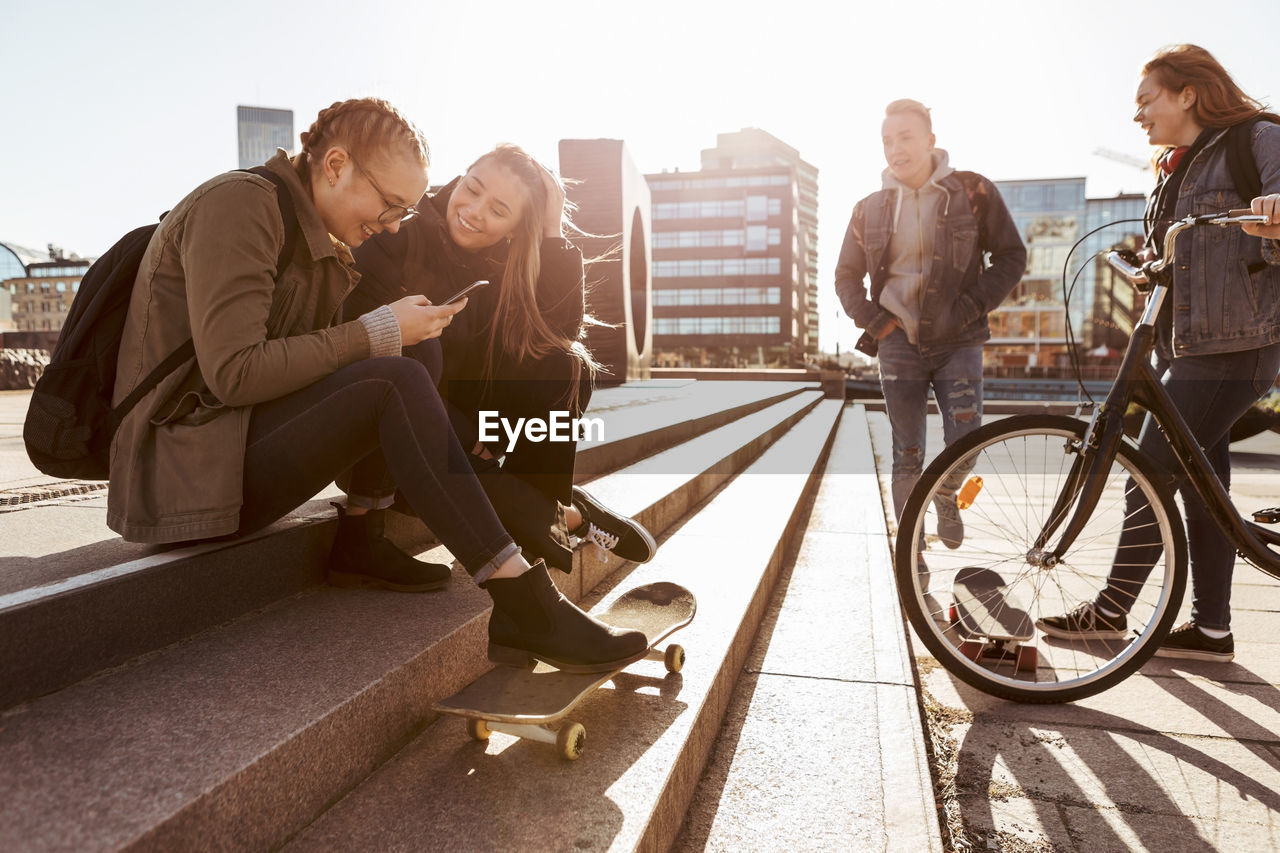 Teenage girls talking on steps while friends standing with bicycle and skateboard in city