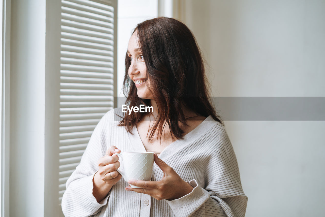 Young woman in cozy knitted cardigan with cup of tea in hands in bright interior at home