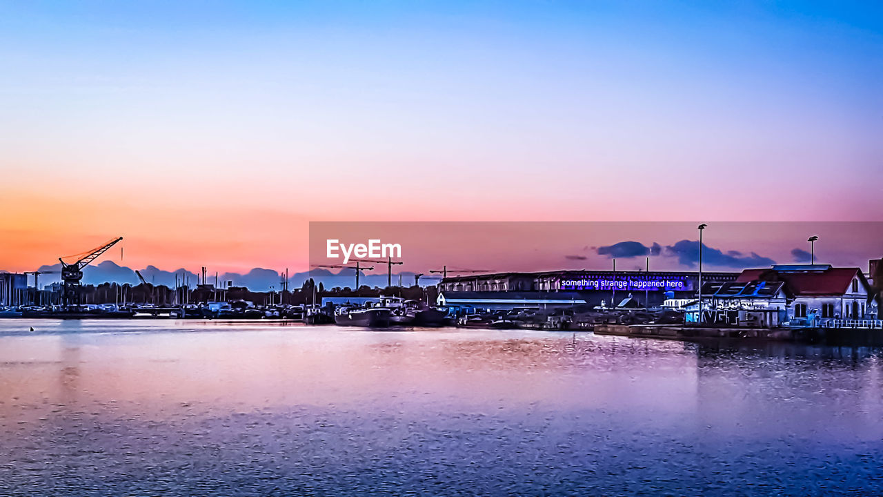 ILLUMINATED BRIDGE OVER SEA AGAINST BUILDINGS AT SUNSET