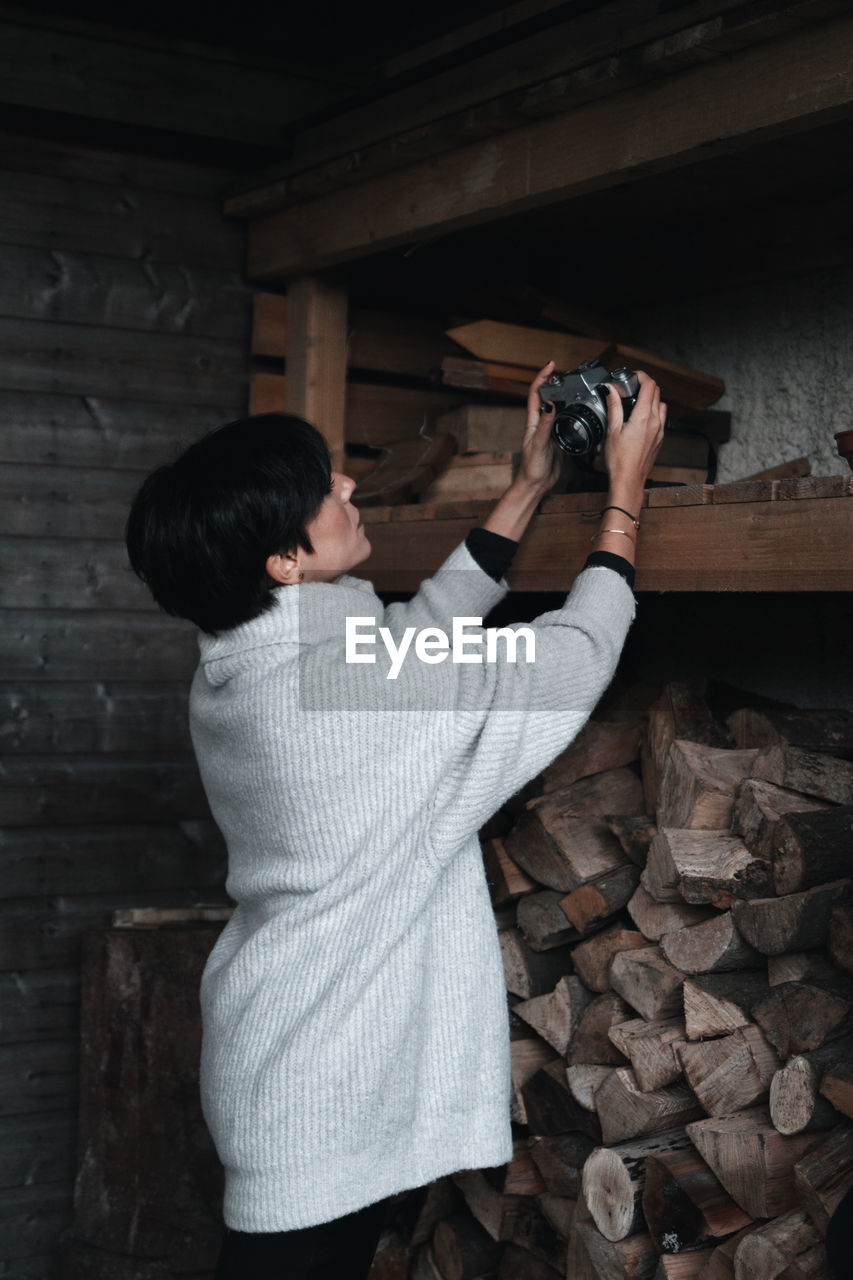 Woman photographing while holding camera on shelf with firewood