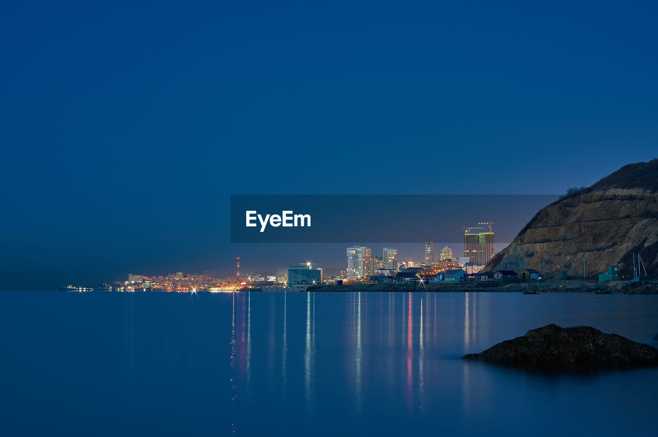 Illuminated city by sea against clear blue sky at night