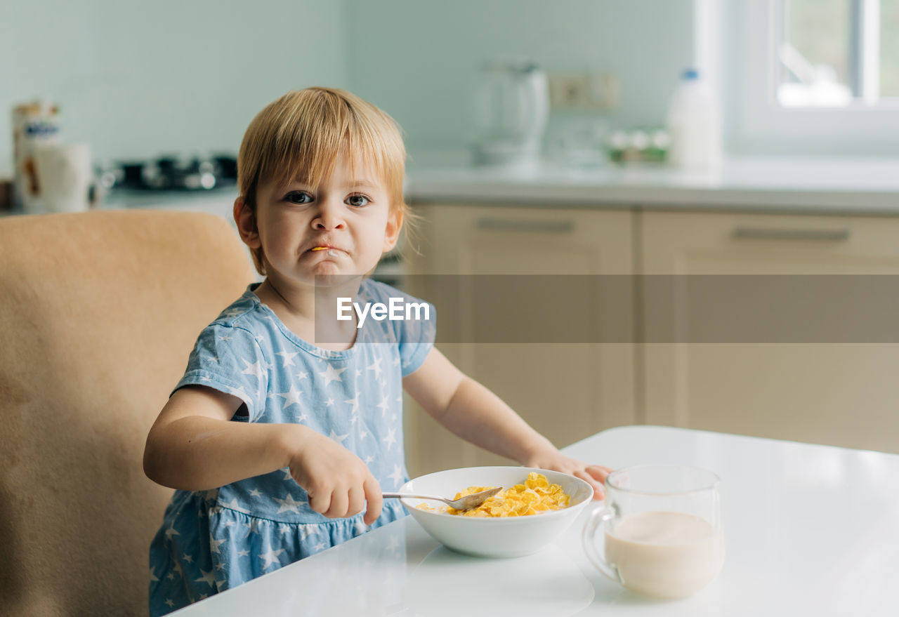 Little toddler girl eats corn flakes with milk in the kitchen in the morning.