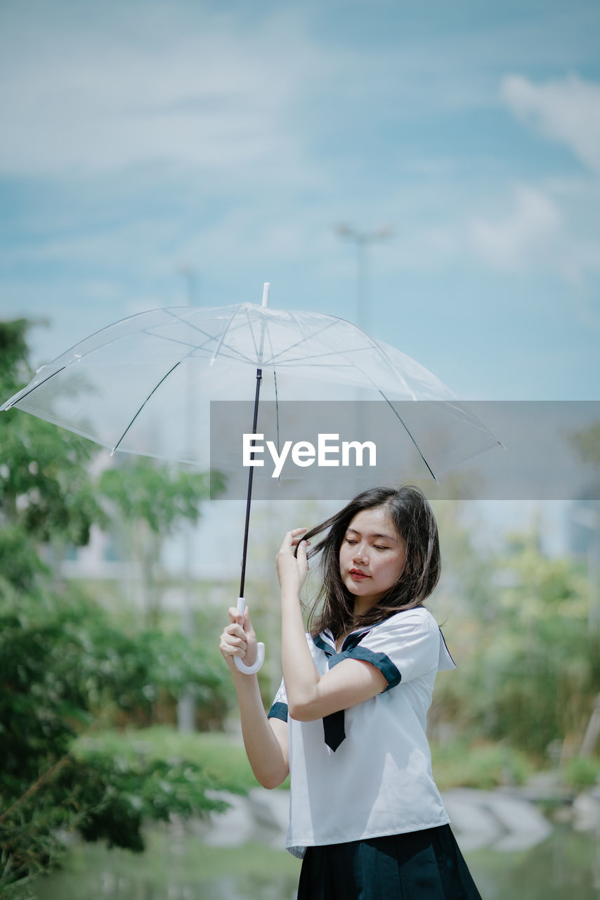 Thoughtful young woman with umbrella standing against sky
