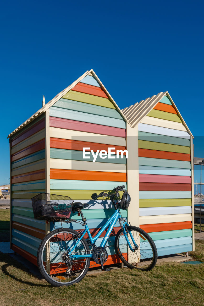 A vibrant beach hut under an azure sky, accompanied by a ready-to-ride bicycle.