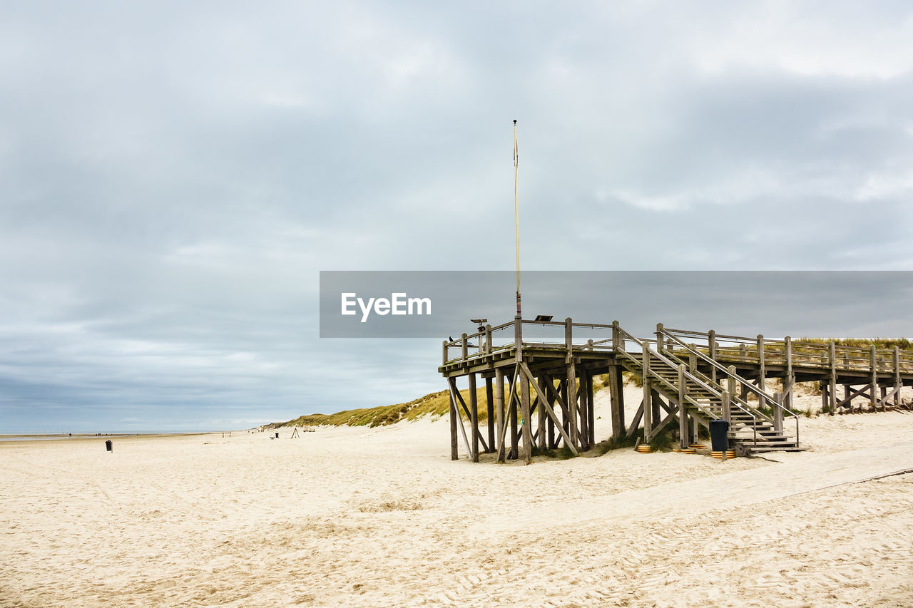 Low angle view of pier on sandy beach against cloudy sky