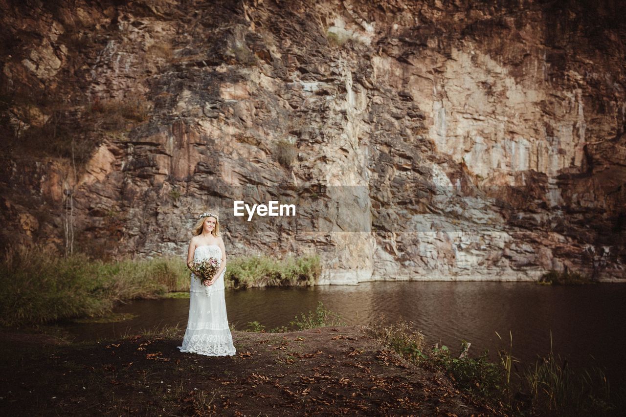 Portrait of woman wearing wedding dress holding bouquet standing by water against rock