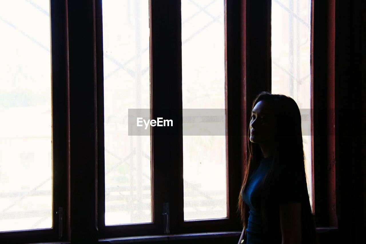 SIDE VIEW OF WOMAN LOOKING THROUGH WINDOW AT HOME