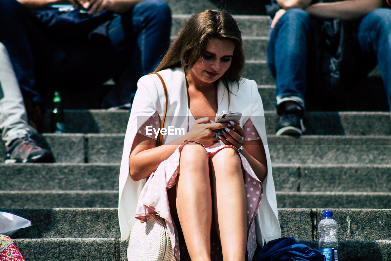 MIDSECTION OF WOMAN USING MOBILE PHONE WHILE SITTING ON STAIRCASE OF PEOPLE