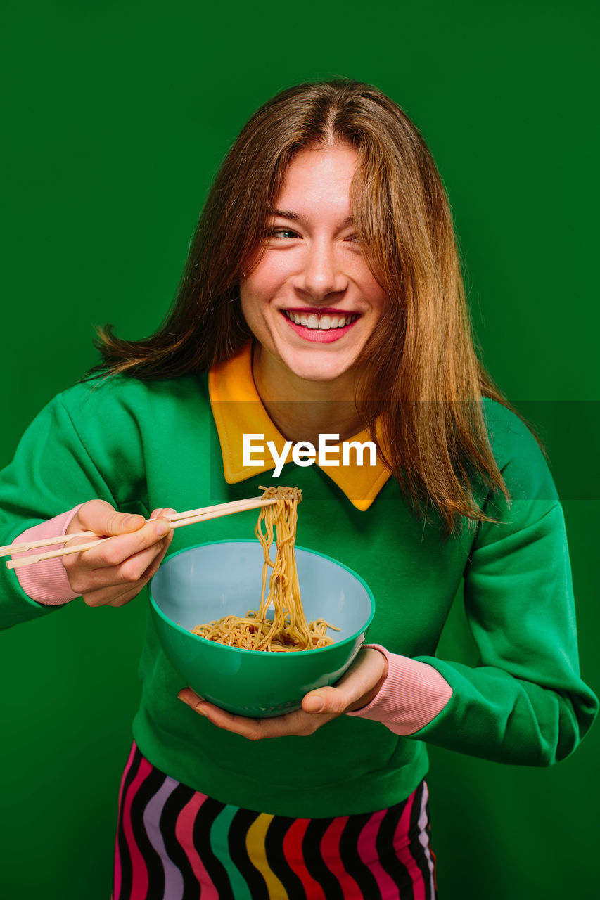 Positive cheerful young female in green shirt looking at camera while eating yummy instant noodles with chopsticks on green background