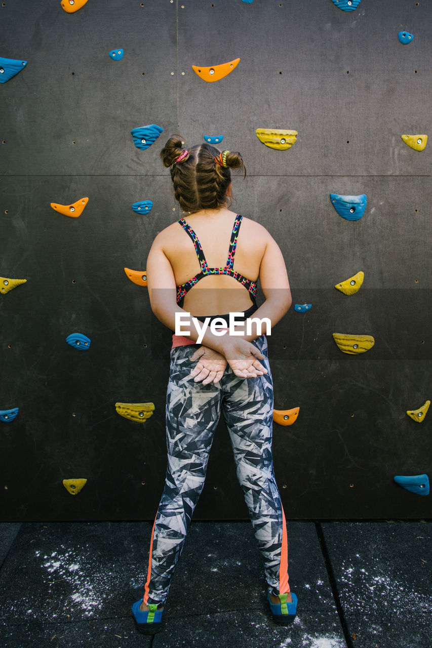Back view of anonymous child in sportswear standing near wall with artificial rocks while training in gym and enjoying weekend