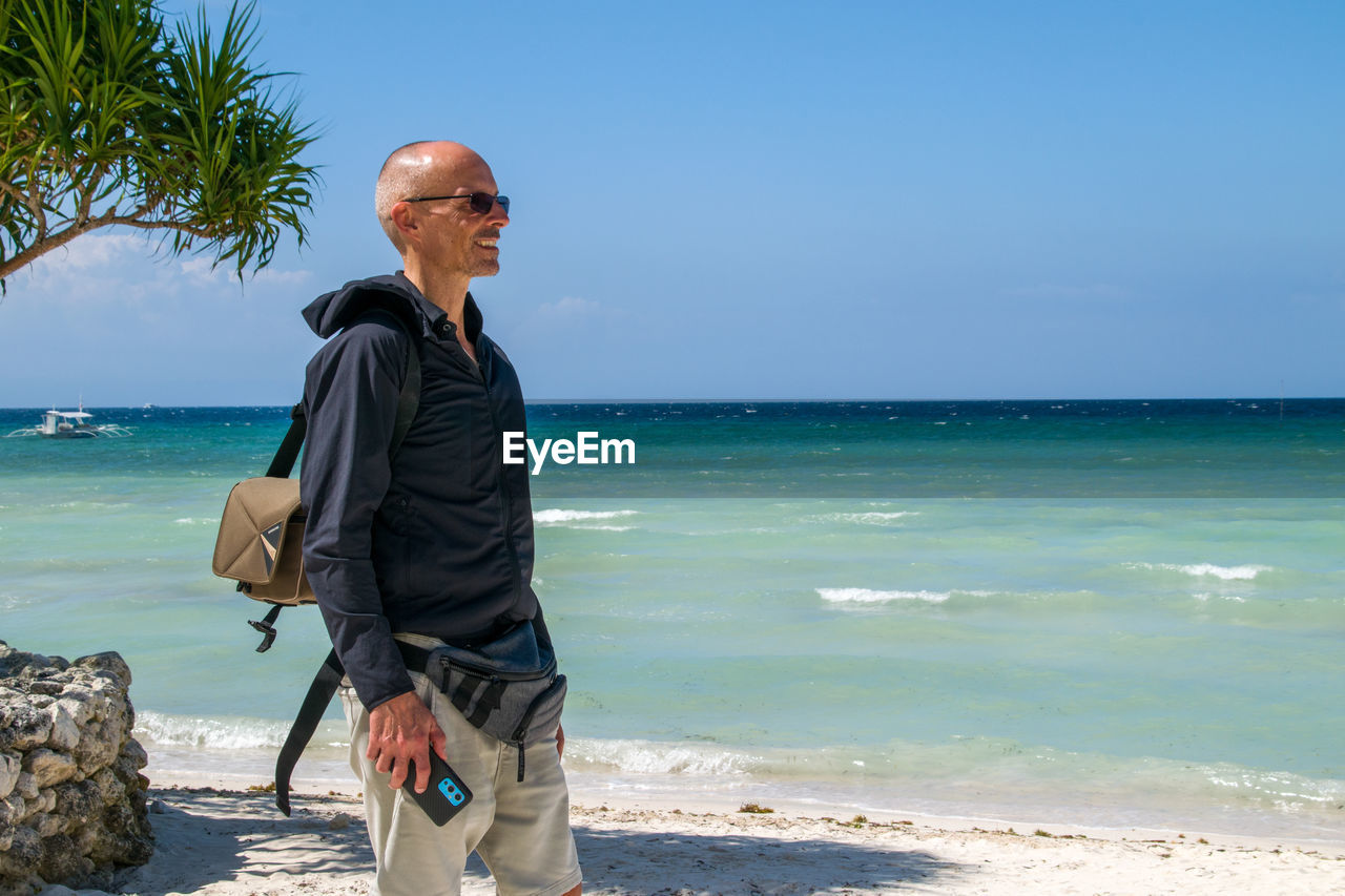 vacation, sea, water, beach, land, one person, shore, ocean, nature, adult, sky, coast, body of water, horizon over water, holiday, trip, senior adult, tropical climate, horizon, men, standing, beauty in nature, day, sunny, scenics - nature, sunlight, leisure activity, travel, clear sky, seniors, blue, travel destinations, sand, bay, relaxation, outdoors, looking, lifestyles, mature adult, tranquility, casual clothing, palm tree, three quarter length, tranquil scene, copy space, person, retirement, full length, idyllic, glasses, clothing, looking away