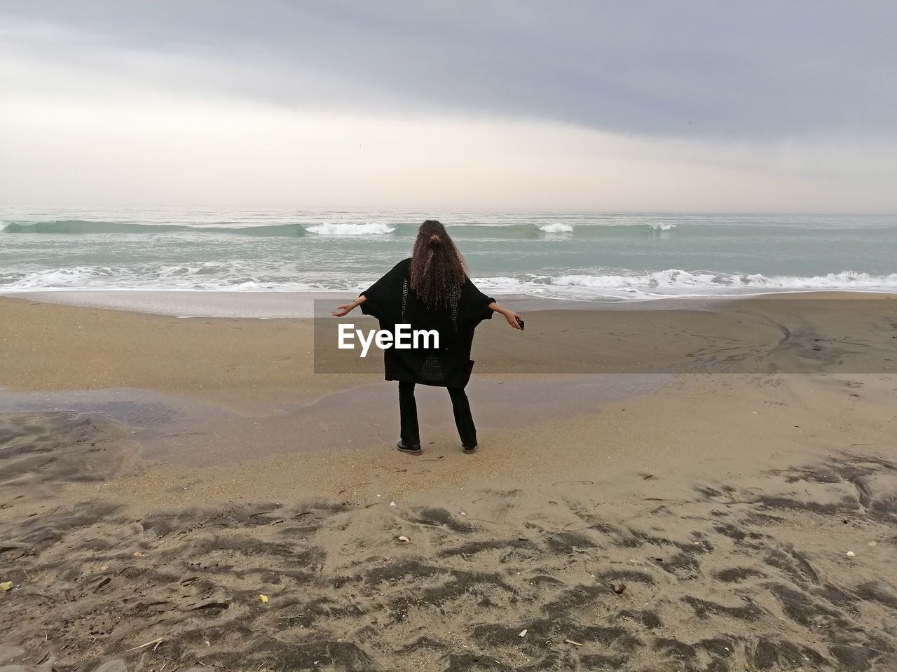 REAR VIEW OF WOMAN STANDING ON BEACH AGAINST SKY