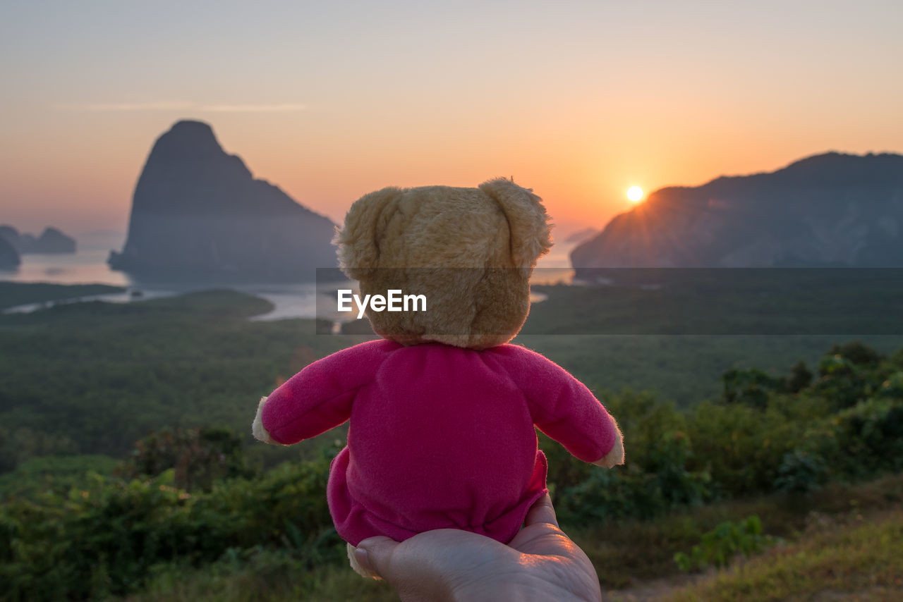 Cropped hand holding teddy bear against sea during sunset