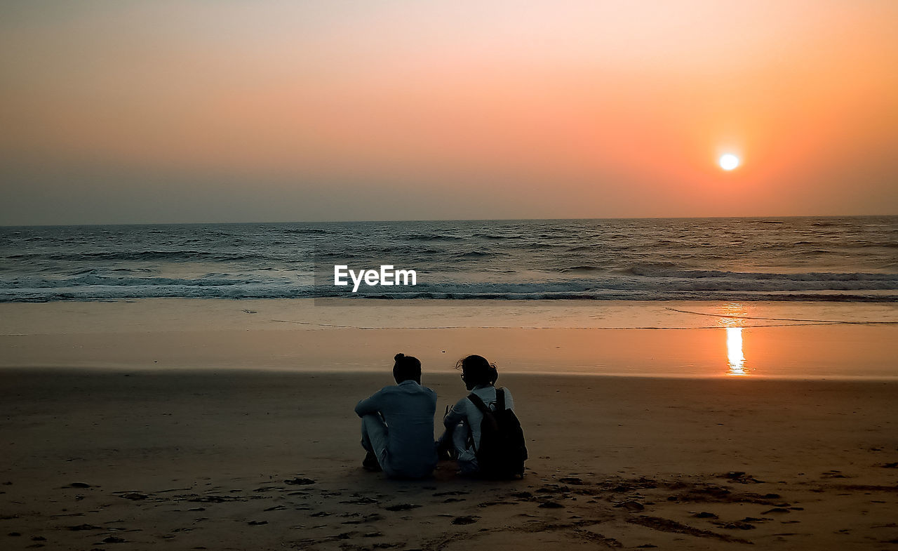 REAR VIEW OF PEOPLE ON BEACH AGAINST SKY DURING SUNSET