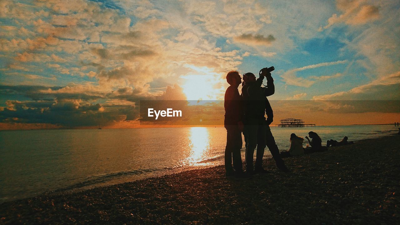 SILHOUETTE PEOPLE STANDING ON BEACH AGAINST ORANGE SKY