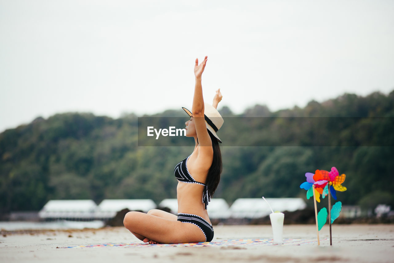 Side view of woman with arms raised sitting at beach against sky