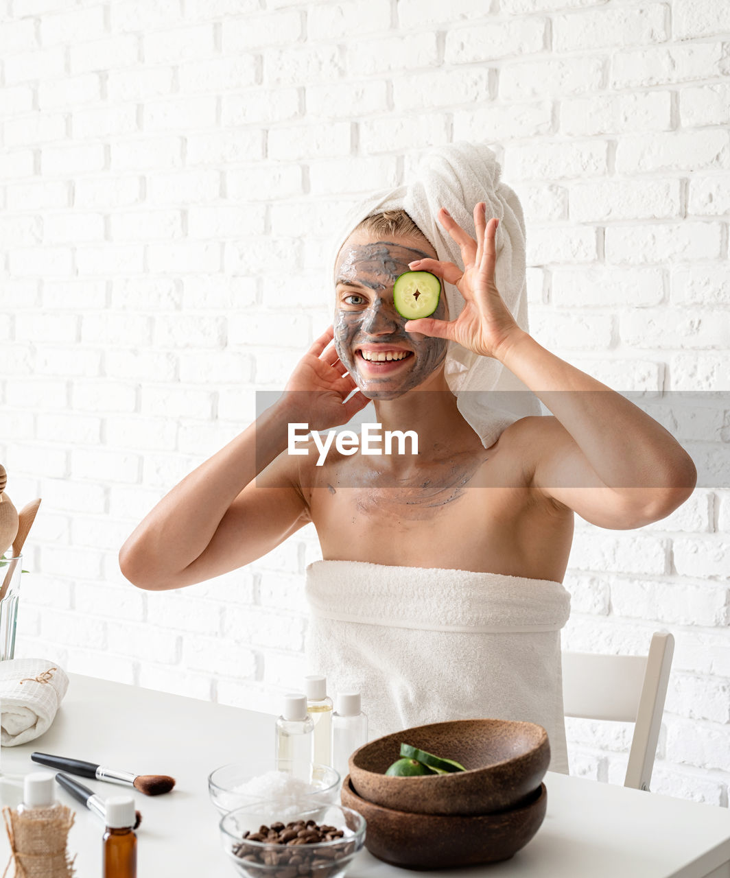 Young woman wearing white towels applying spa facial mask on her face with a cosmetic brush