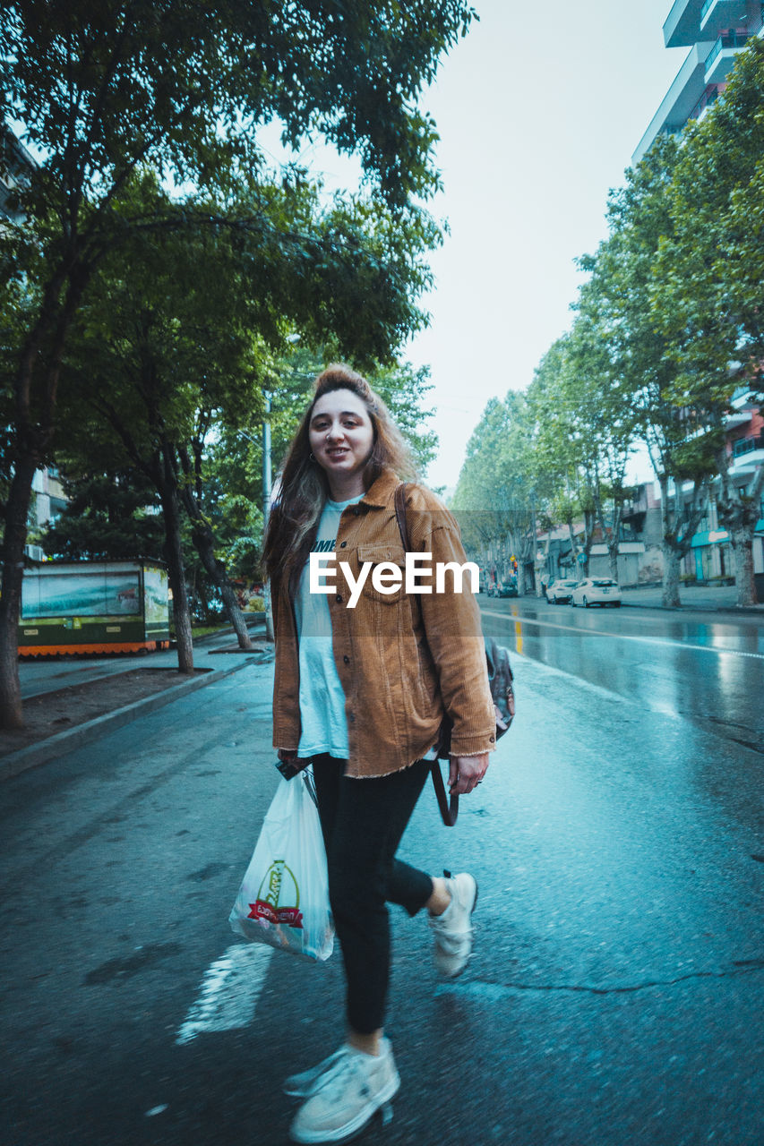 PORTRAIT OF YOUNG WOMAN STANDING ON ROAD IN CITY