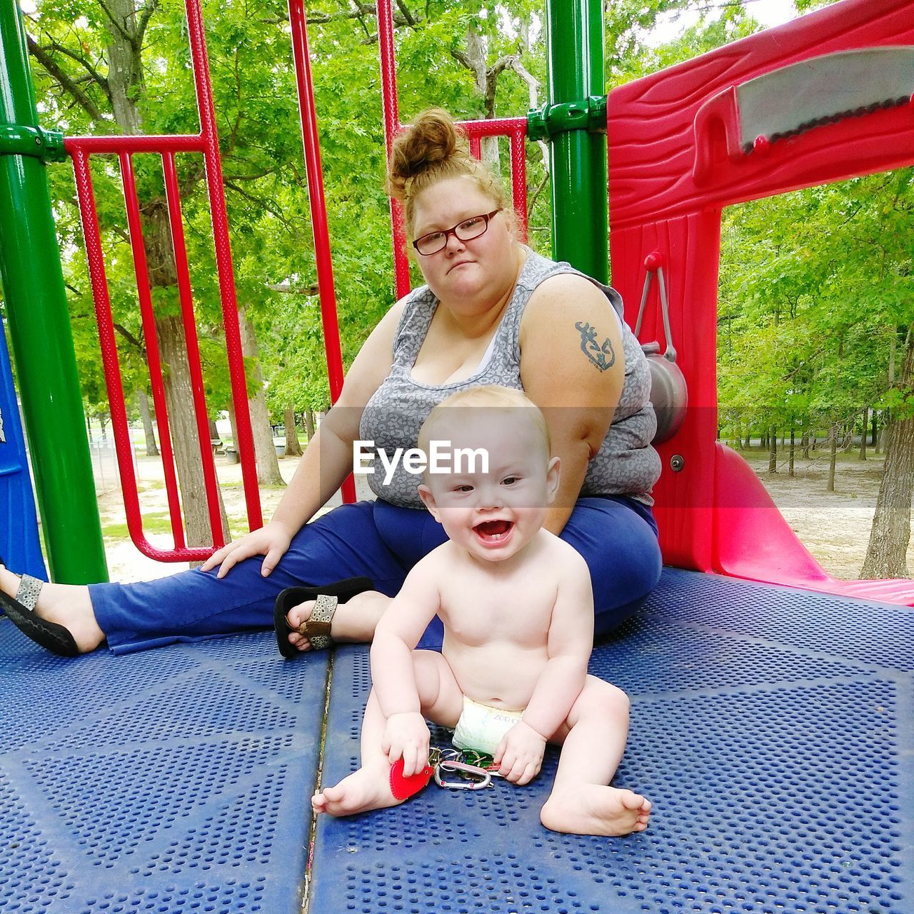 Portrait of woman with shirtless baby sitting on outdoor play equipment at playground