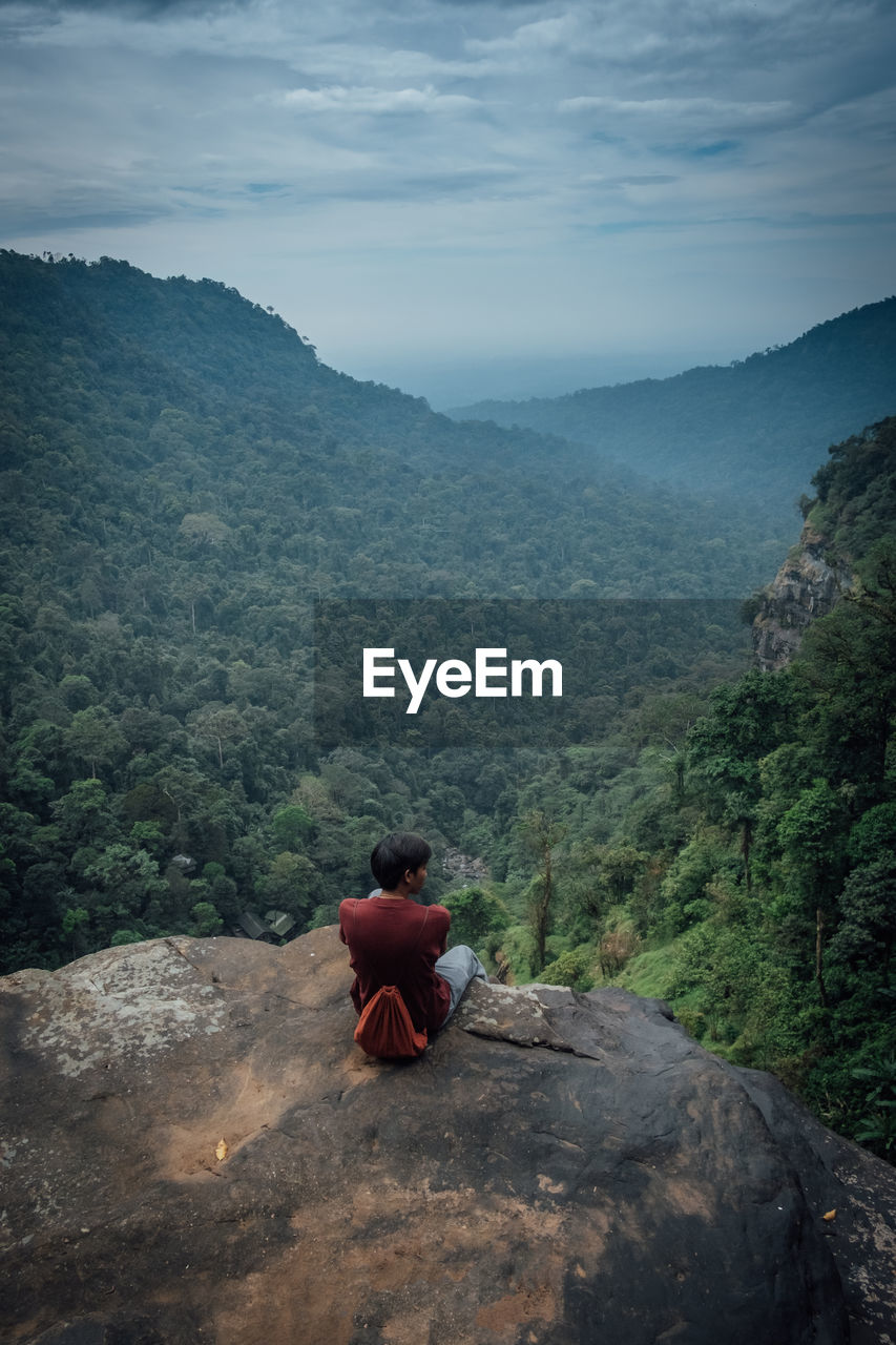 REAR VIEW OF MAN SITTING ON LANDSCAPE AGAINST MOUNTAINS