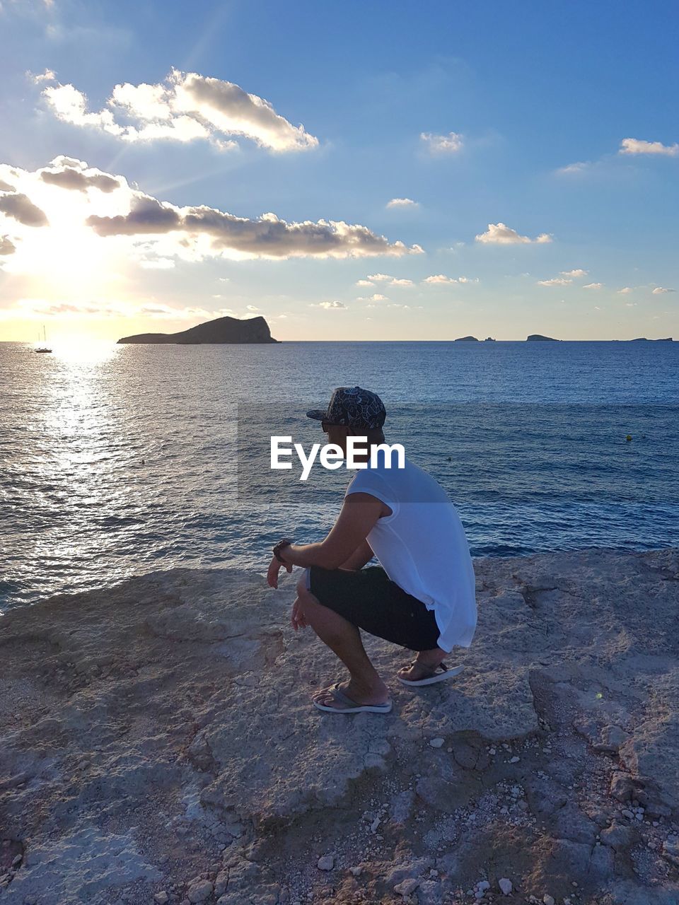 Side view of man crouching on rock at beach during sunset