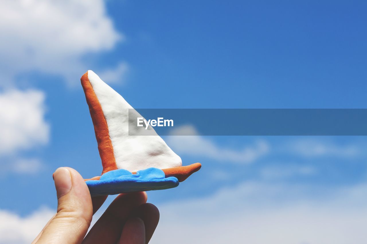 Fingers are holding a plasticine sailboat with a white sail against a blue sky with white clouds