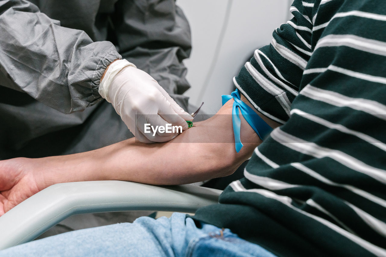 Professional female medical specialist in protective uniform preparing arm of anonymous patient for blood test in modern lab