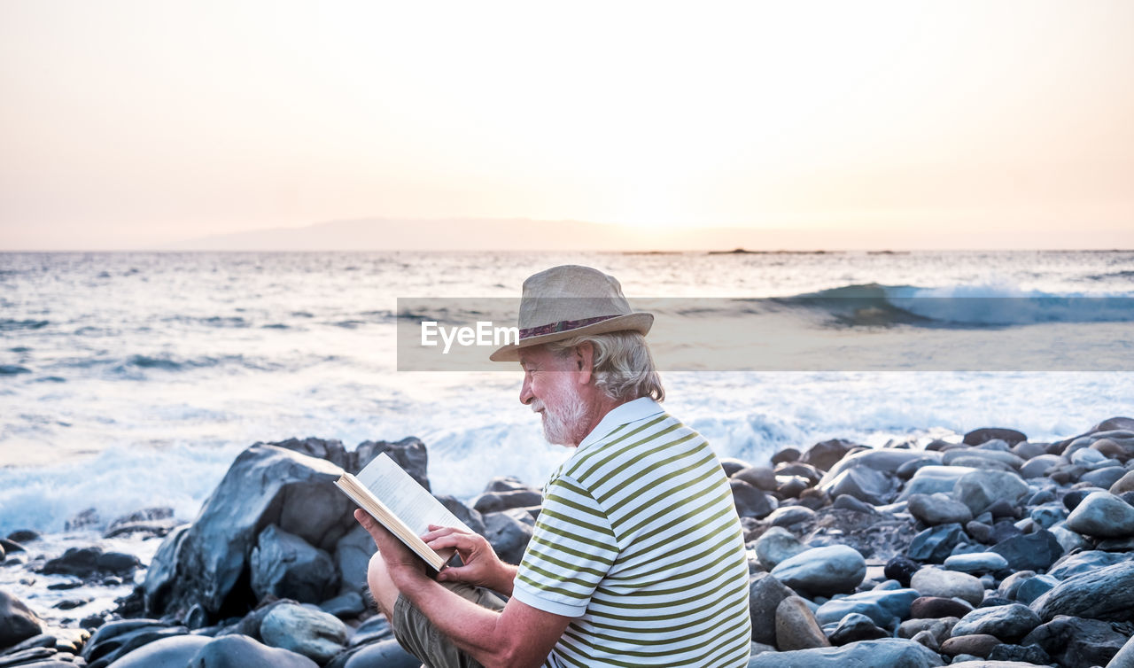 Senior man reading book while sitting on rock at beach during sunset