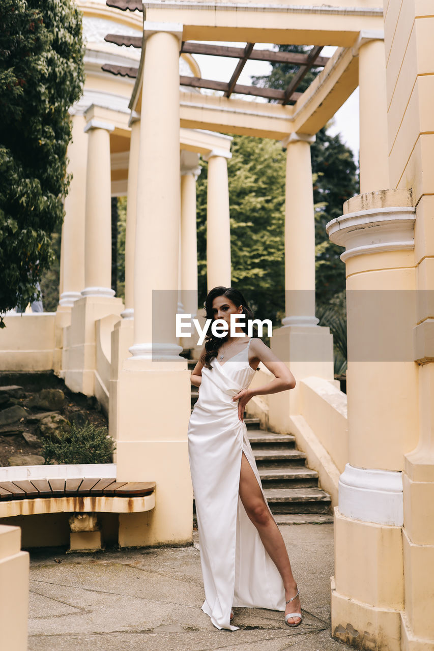 A beautiful brunette lady in an elegant wedding dress poses among the columns in the old city park