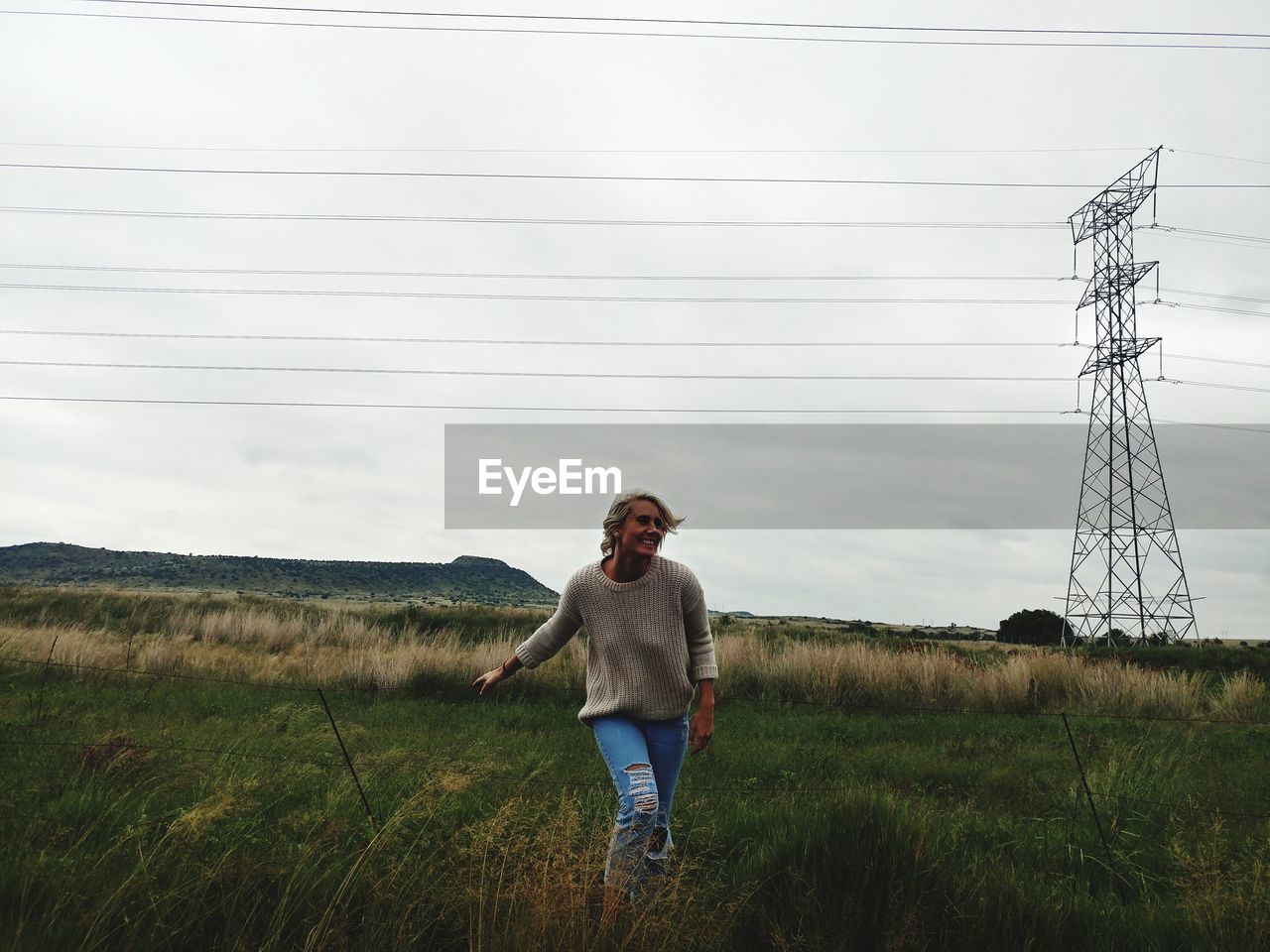 Smiling woman standing on field against electricity pylon