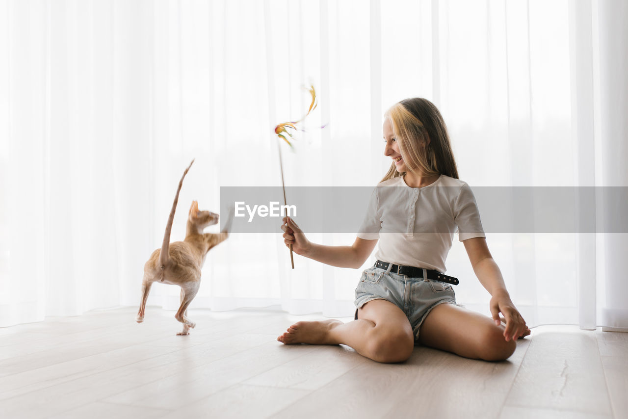 Caucasian teen girl sitting on the floor playing with a cat cornish rex with a stick teaser