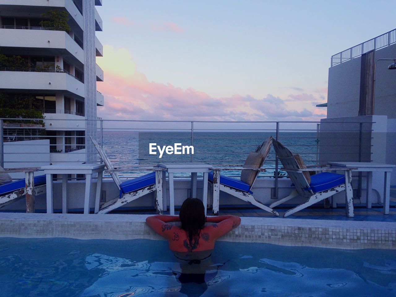Rear view of woman relaxing in swimming pool in front of sea at dusk