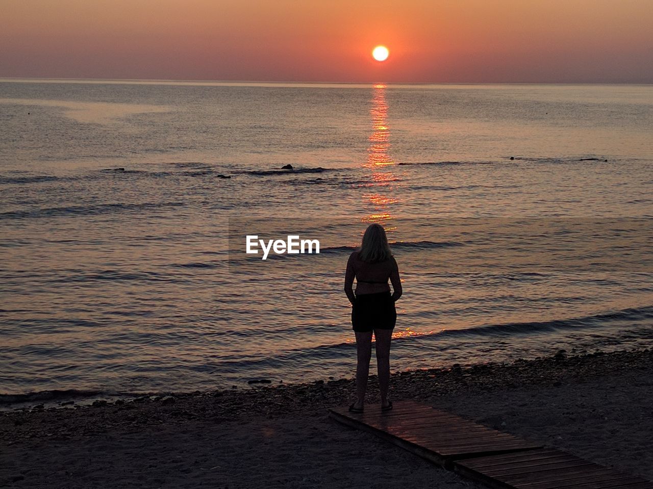 Full length rear view of woman standing at beach against sky during sunset