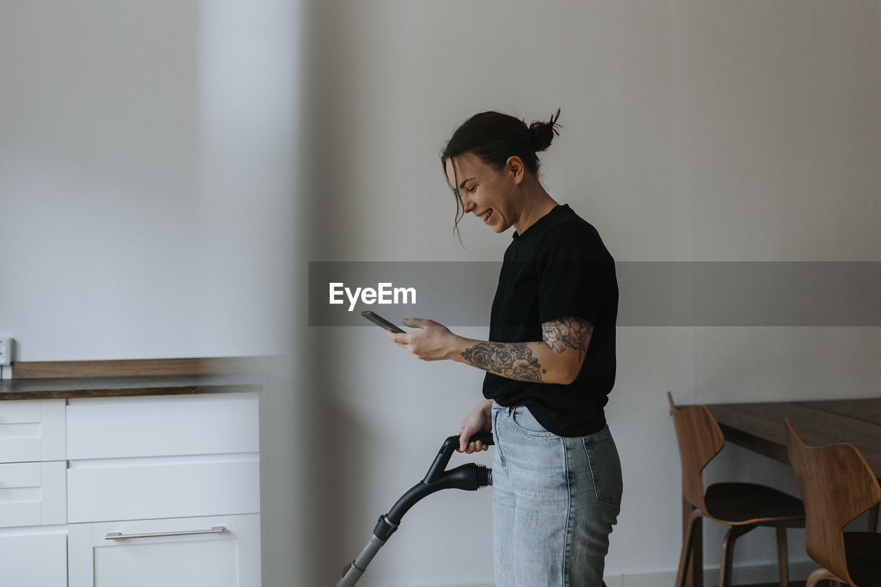 Smiling woman using cell phone while vacuum cleaning room