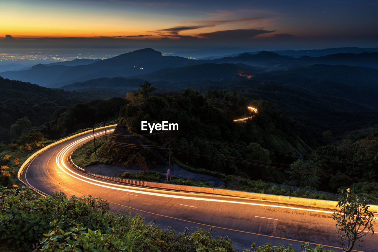LIGHT TRAILS ON ROAD BY MOUNTAINS AGAINST SKY AT SUNSET