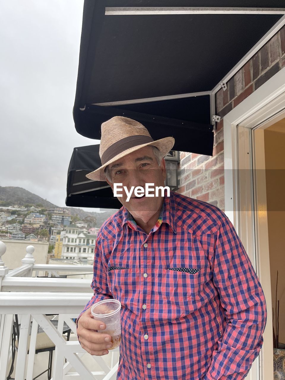 Portrait of man wearing hat holding drink standing against built structure on balcony 