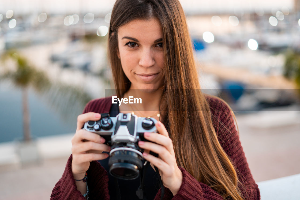 PORTRAIT OF SMILING YOUNG WOMAN HOLDING CAMERA