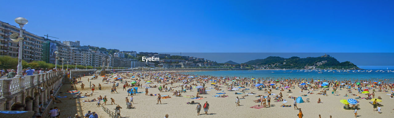beach, land, crowd, water, sky, sea, body of water, large group of people, vacation, holiday, trip, group of people, nature, sand, travel destinations, coast, travel, clear sky, architecture, blue, tourism, summer, leisure activity, walkway, sunny, lifestyles, sports, coastline, relaxation, enjoyment, outdoors, tourist, day, ocean, panoramic, shore, scenics - nature, city, built structure, men, adult, sunlight