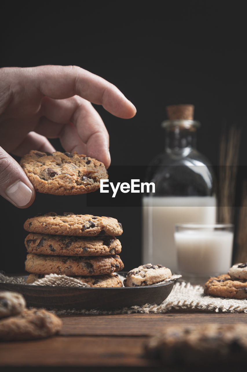 Hand holding chocolate chip cookie with glass and a bottle of milk on wooden base, dark background,.