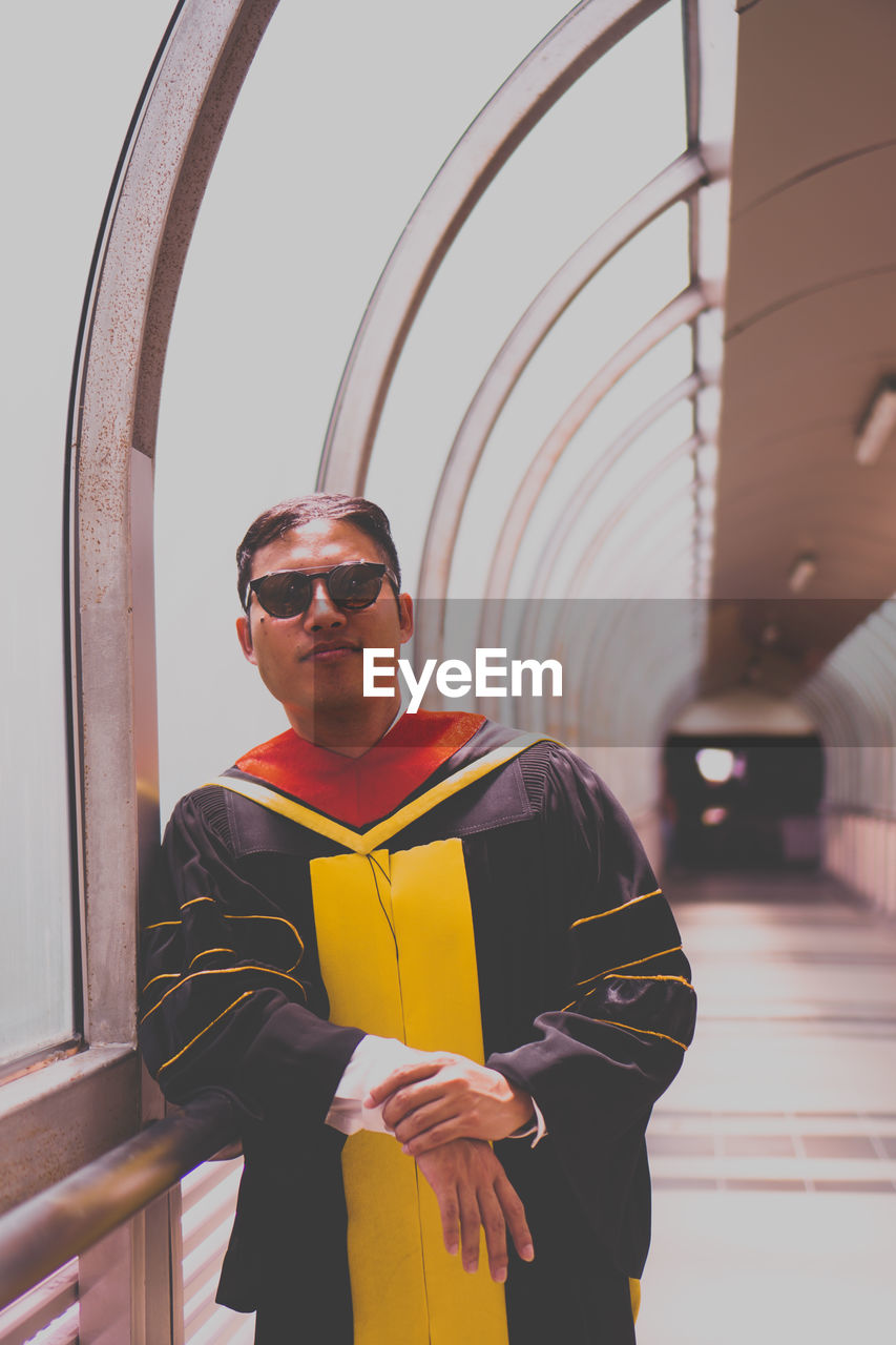 Portrait of young man wearing sunglasses and graduation gown while standing in corridor