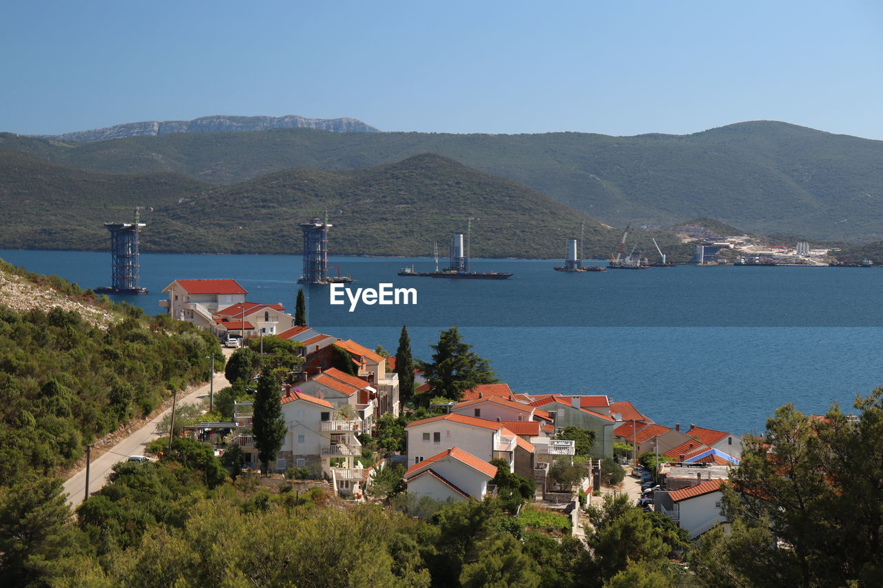 HIGH ANGLE VIEW OF TOWNSCAPE BY SEA AND MOUNTAINS AGAINST SKY