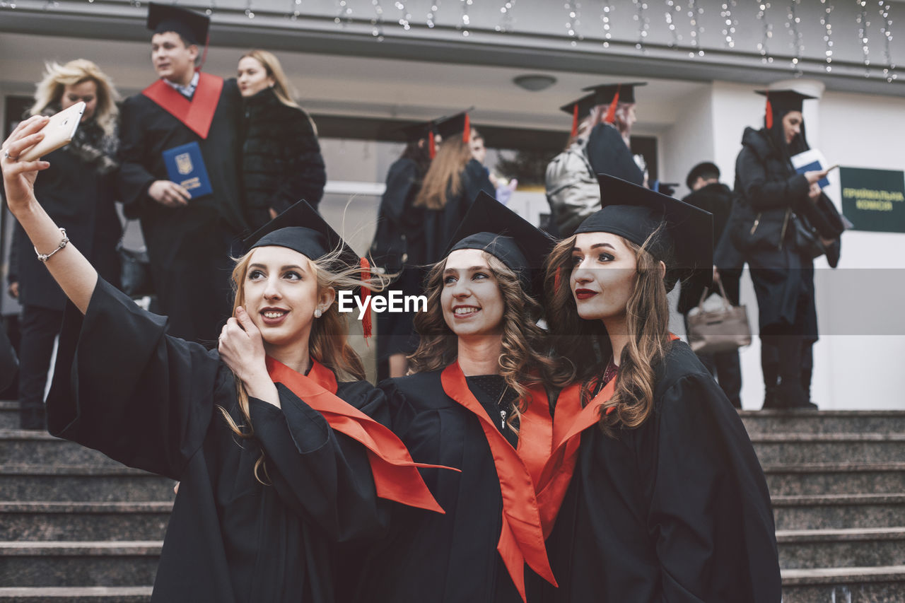 Happy female friends wearing graduation gowns taking selfie while standing on steps