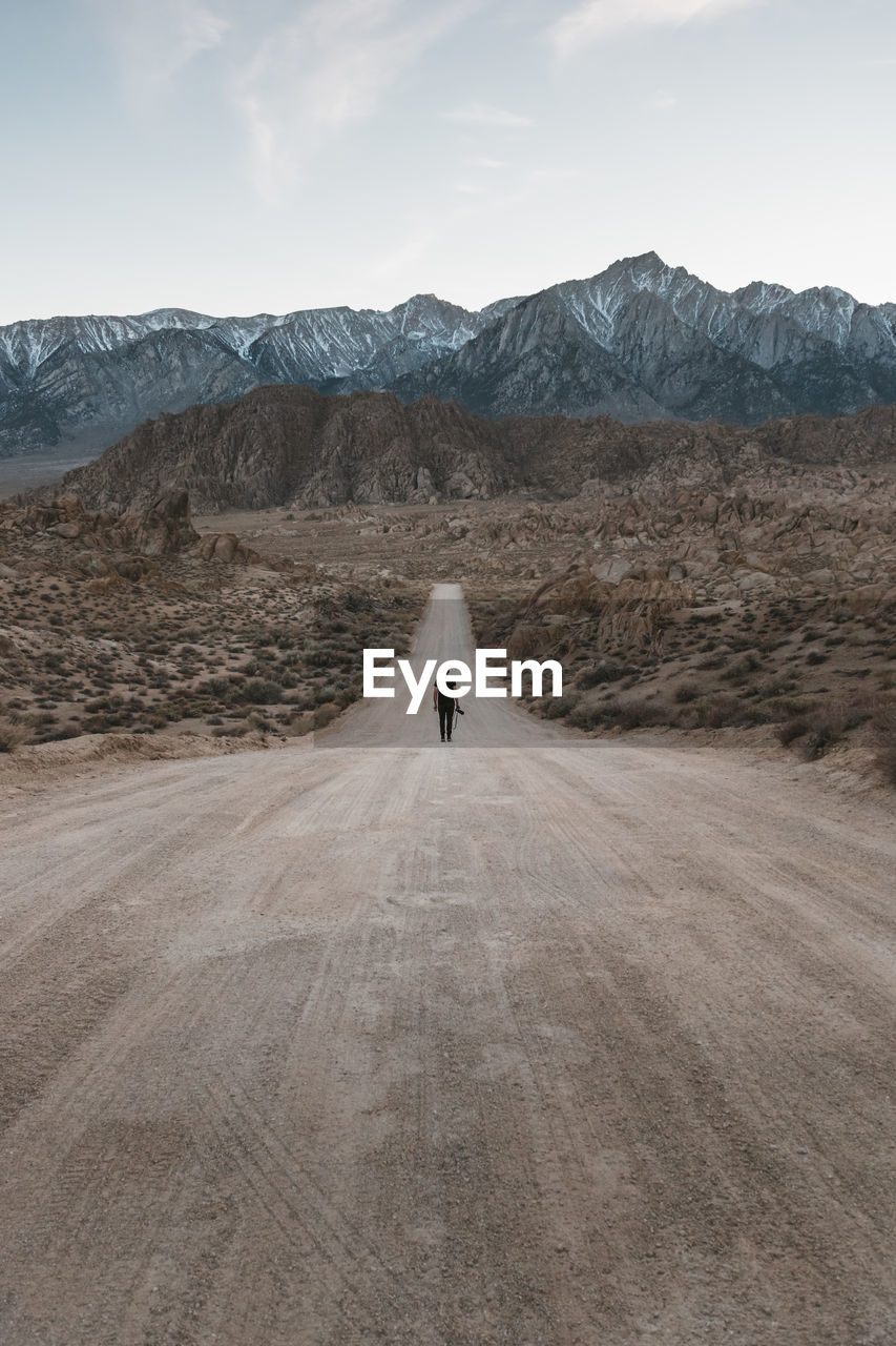 Rear view of man walking on dirt road against sky