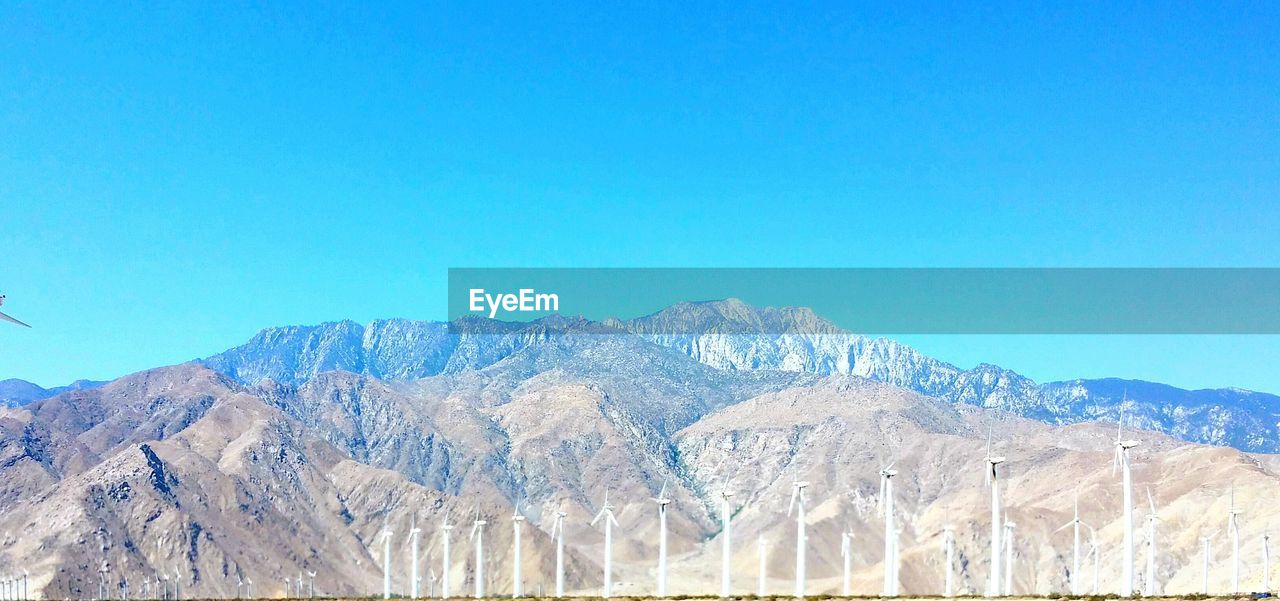 Wind turbines by mountains against clear blue sky