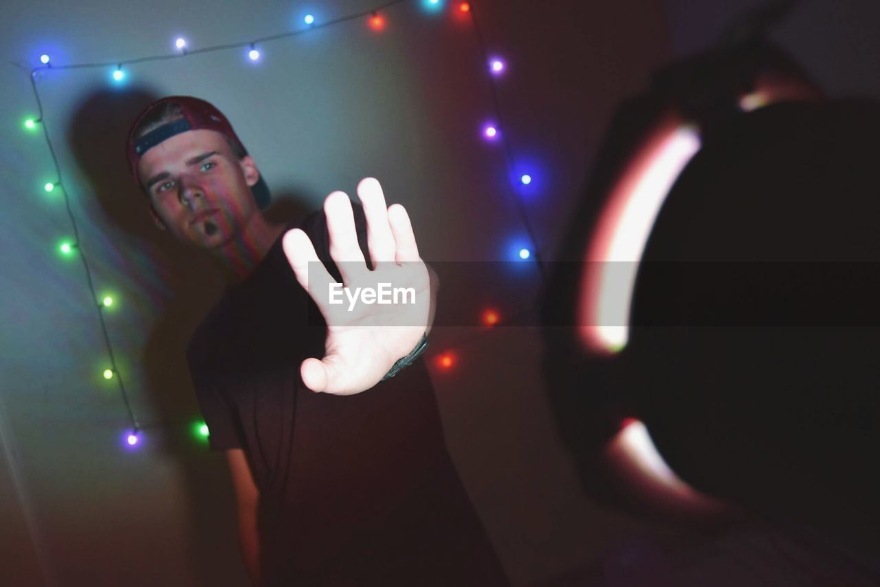 Young man gesturing at illuminated halogen light while standing against decoration on wall