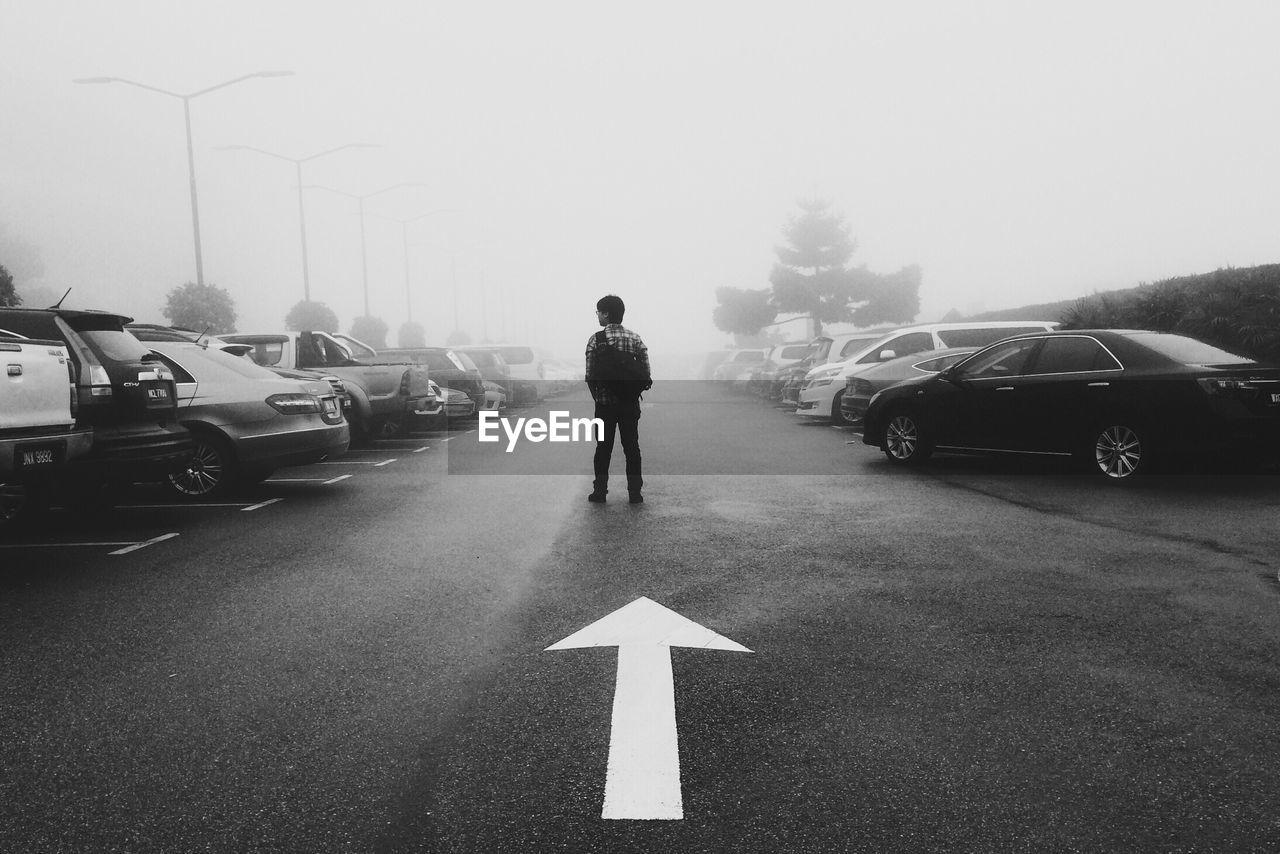 Rear view of man standing by arrow symbol amidst cars during foggy weather