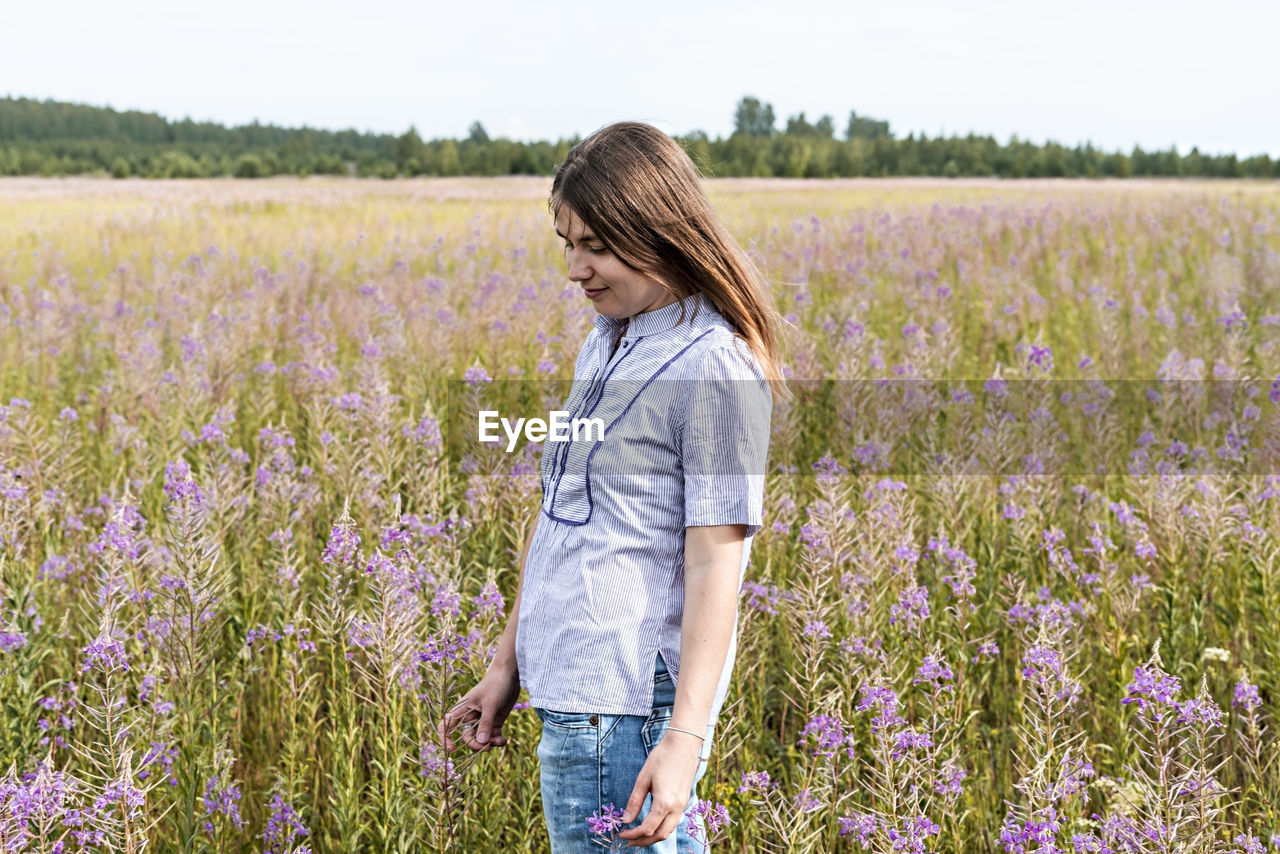 Young beautiful smiling blond woman in purple shirt walking in the meadow among flowers of fireweed