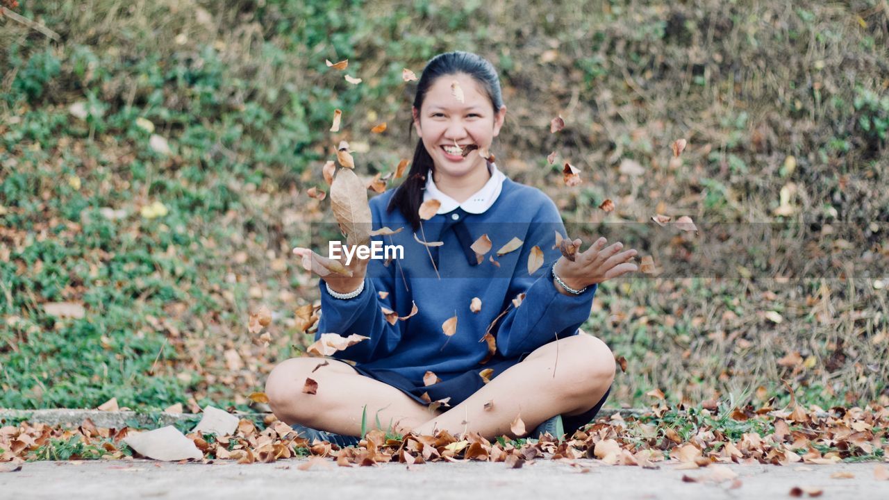 Portrait of smiling young woman throwing leaves while sitting on field in park