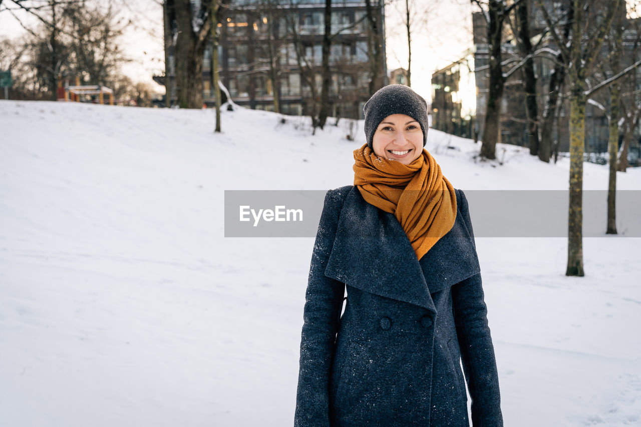 Smiling woman in a winter coat, scarf and hat against the backdrop of a winter snowy city